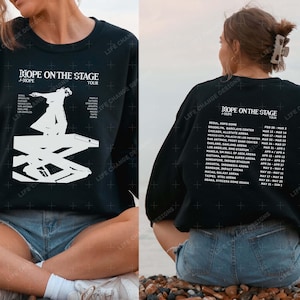 May include: Black sweatshirt with white text that reads "Hope on the Stage" and "J-Hope Tour". The sweatshirt features a graphic of a person on a stage with a list of cities and dates for the tour.