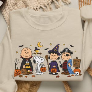 Könnte beinhalten: Ein cremefarbenes Sweatshirt mit Rundhalsausschnitt und einem Grafikdruck der Peanuts-Figuren in Halloween-Kostümen, darunter ein Vampir, eine Mumie, eine Hexe und ein Pirat, umgeben von Kürbissen, Fledermäusen und einem Schild mit der Aufschrift Trick or Treat.