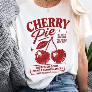 Könnte beinhalten: Ein weißes T-Shirt mit einer roten Grafik von zwei Kirschen und dem Text "CHERRY PIE", "OH SH*T GRAHAM YOU KNOW THIS ONE?" sowie "TASTES SO GOOD, MAKE A GROWN MAN CRY SWEET CHERRY PIE-EEEEEEEE" in einer kräftigen, vintage-inspirierten Schrift.