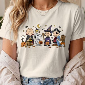 May include: A cream-coloured short-sleeve t-shirt featuring a graphic print of Peanuts characters dressed in Halloween costumes, including a vampire, a mummy, a witch, and a pirate, with pumpkins, bats, and a sign that reads Trick or Treat.