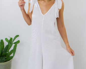 May include: A white linen jumpsuit with a V-neck and tie straps. The jumpsuit has a relaxed fit and a flowing silhouette.