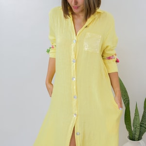 May include: A yellow linen dress with a floral print hem and a sequined pocket. The dress has a button-up front and long sleeves.