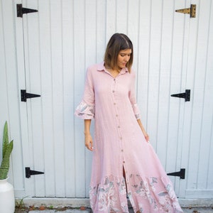 May include: A woman wearing a long, pink, button-down dress with a floral print. The dress has a ruffled hem and 3/4 sleeves. She is standing in front of a white wooden door.