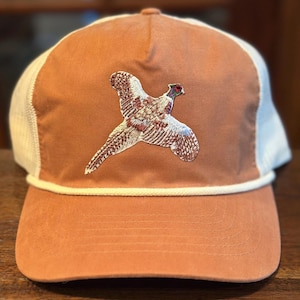 May include: A rust-colored baseball cap with a white mesh back and a white rope detail. The cap features an embroidered pheasant in flight, showcasing detailed feather patterns in shades of brown, white, and red.