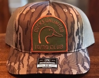 May include: A camouflage trucker hat with a Ducks Unlimited 10/30 Club patch and a Richardson 112 adjustable label.
