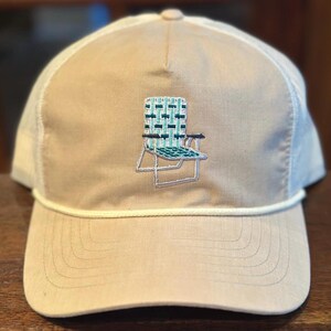 May include: Tan and white trucker hat with a cream-colored rope detail. The hat features an embroidered image of a beach chair in shades of teal and black. The hat has a mesh back and a curved brim.