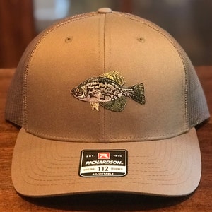 May include: A tan Richardson 112 Trucker hat with a mesh back and an embroidered fish design. The hat features a curved brim and an adjustable strap. The fish is detailed with shades of green, black, and tan.