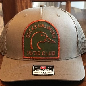 May include: A gray and brown mesh trucker hat with a Ducks Unlimited 10/30 Club patch. The patch features a green duck head with an orange outline. The hat is adjustable and has a Richardson 112 label.