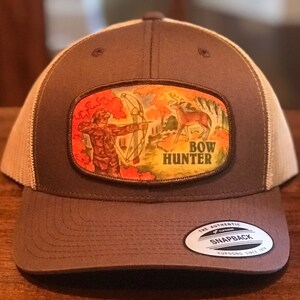 May include: Brown and tan trucker hat with a patch featuring a bow hunter aiming at a deer in a forest scene. The patch has the words "BOW HUNTER" in yellow. The hat has a snapback closure and a Yupoong tag.