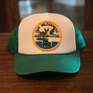 May include: White and green trucker hat with a patch that says "Our Wilderness" and features a deer, a bird, and a fish.