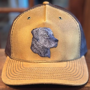 May include: A tan and brown baseball cap featuring a black dog portrait on the front. The cap has a mesh back, a curved brim, and two small eyelets on the crown. The dog portrait is embroidered.