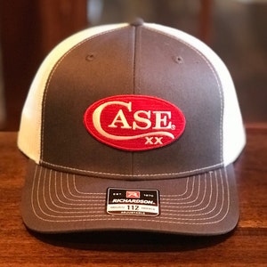 May include: A brown and white baseball cap with a red oval patch that says "CASE XX" on it. The cap is made by Richardson and is adjustable.