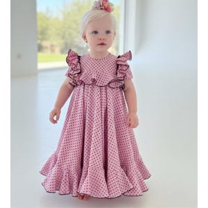 Puede incluir: Un vestido rosa con lunares negros, ribetes negros y mangas con volantes. El vestido tiene una falda acampanada y lo lleva una niña pequeña. Una diadema floral adorna la cabeza de la niña. El vestido es de color rosa claro con lunares negros.