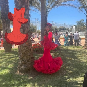 Puede incluir: Una mujer con un vibrante vestido de flamenco rojo con mangas con volantes y una falda acampanada, posando al aire libre. El vestido presenta un corpiño ajustado y una falda larga y fluida. Un tocado floral rojo complementa el vestido. El fondo incluye palmeras y una fuente.