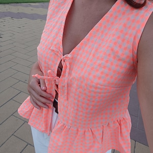 Puede incluir: Una blusa sin mangas de cuadros naranja y blanco con un dobladillo con volantes y un lazo en el escote.