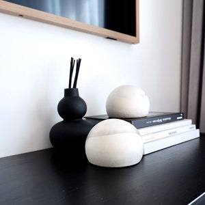 Può includere: Tre sfere di ceramica bianca, di cui due sono su una pila di libri con i titoli "BAHAUS" e "MONOCHROME" visibili. La terza sfera è su una superficie nera accanto a un vaso di ceramica nero con bastoncini neri.