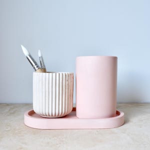 May include: A pink ceramic tray with two pink ceramic containers. One container is taller and the other is shorter and has a white ribbed design. There are three paintbrushes in the shorter container.