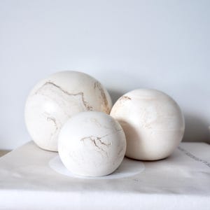Pode incluir: Três esferas decorativas brancas de tamanhos variados com veios castanhos sutis. As esferas estão dispostas sobre uma superfície branca, criando uma estética minimalista. A esfera maior tem cerca de 15 cm de diâmetro.