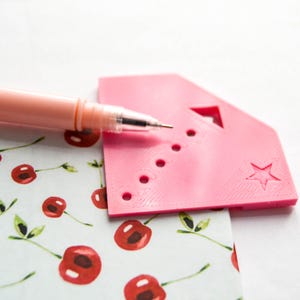 Puede incluir: Una herramienta de plástico rosa con una estrella y una fila de agujeros. La herramienta está sobre una hoja de papel con un patrón de cerezas.