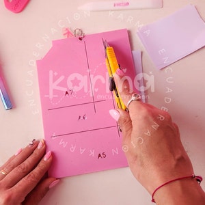 May include: A pink cutting mat with grid lines and the text "Karma Encuadernaion" printed on it. A person is using a yellow utility knife to cut along a line on the mat.
