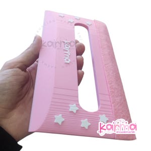 Puede incluir: Una herramienta de espátula de plástico rosa claro para encuadernación con un recorte rectangular, adornos en forma de estrella blanca y un borde de fieltro texturizado. La palabra karma está grabada en letras blancas en el centro.