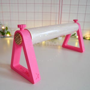 May include: A pink plastic craft tool featuring two triangular side supports connected by a horizontal white cylindrical rod. The rod is secured at each end by pink adjustable knobs. A small wooden circular accent is visible on the side support.