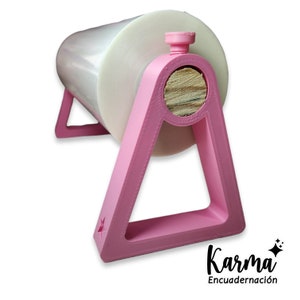 Pode incluir: Suporte de plástico rosa com uma cavilha de madeira para segurar um rolo de filme plástico transparente. O suporte tem uma forma de estrela na parte inferior e o texto "Karma Encuadernación" é visível abaixo do suporte.