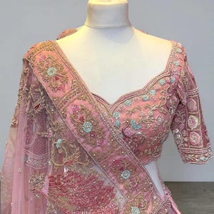 May include: A light pink embroidered blouse and dupatta set. The blouse features intricate floral and peacock designs with gold thread and light blue accents. The dupatta has a matching border and a sheer, delicate appearance.
