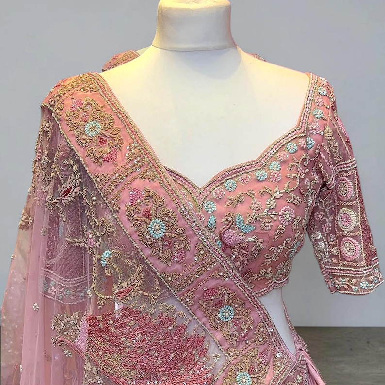 May include: A light pink embroidered blouse and dupatta set. The blouse features intricate floral and peacock designs with gold thread and light blue accents. The dupatta has a matching border and a sheer, delicate appearance.