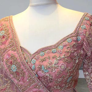 May include: A pink embroidered blouse with floral and peacock motifs. The V-neck blouse has long sleeves and is adorned with detailed beadwork and sequins. The fabric is a sheer, delicate material, with a detailed trim around the neckline.