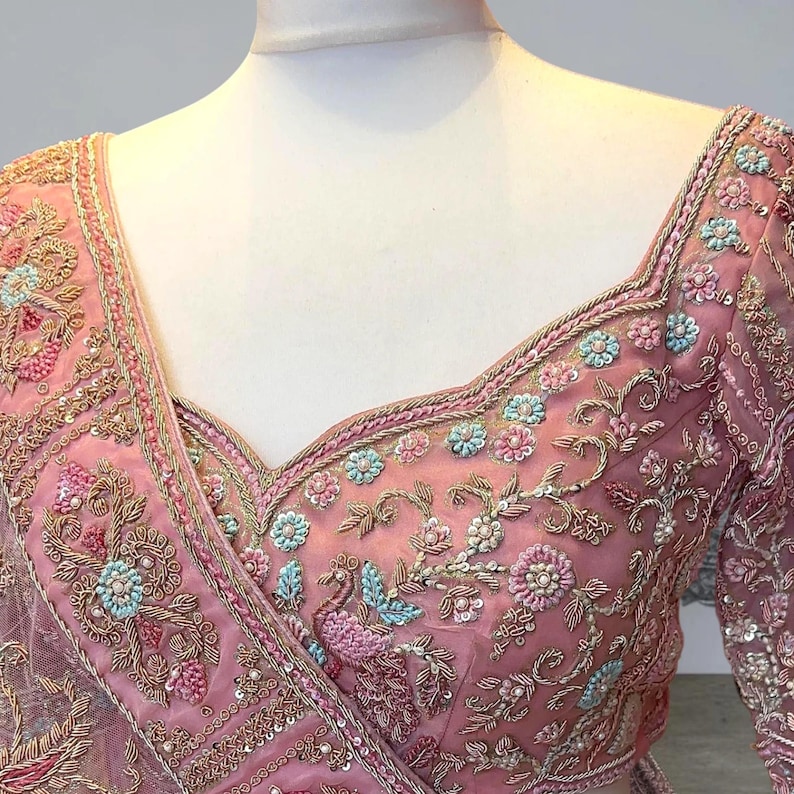 May include: A pink embroidered blouse with floral and peacock motifs. The V-neck blouse has long sleeves and is adorned with detailed beadwork and sequins. The fabric is a sheer, delicate material, with a detailed trim around the neckline.