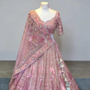 May include: A pink embroidered lehenga choli set on a mannequin. The set comprises a top with short sleeves, a long skirt, and a sheer dupatta. The outfit features detailed embroidery with floral and peacock motifs, and decorative tassels.