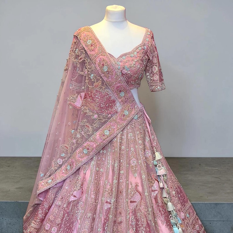 May include: A pink embroidered lehenga choli set on a mannequin. The set comprises a top with short sleeves, a long skirt, and a sheer dupatta. The outfit features detailed embroidery with floral and peacock motifs, and decorative tassels.