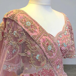 May include: A pink embroidered garment with intricate floral and leaf patterns. The garment features gold thread detailing, sequins, and a sheer overlay. The design includes a decorative border and is displayed on a mannequin.