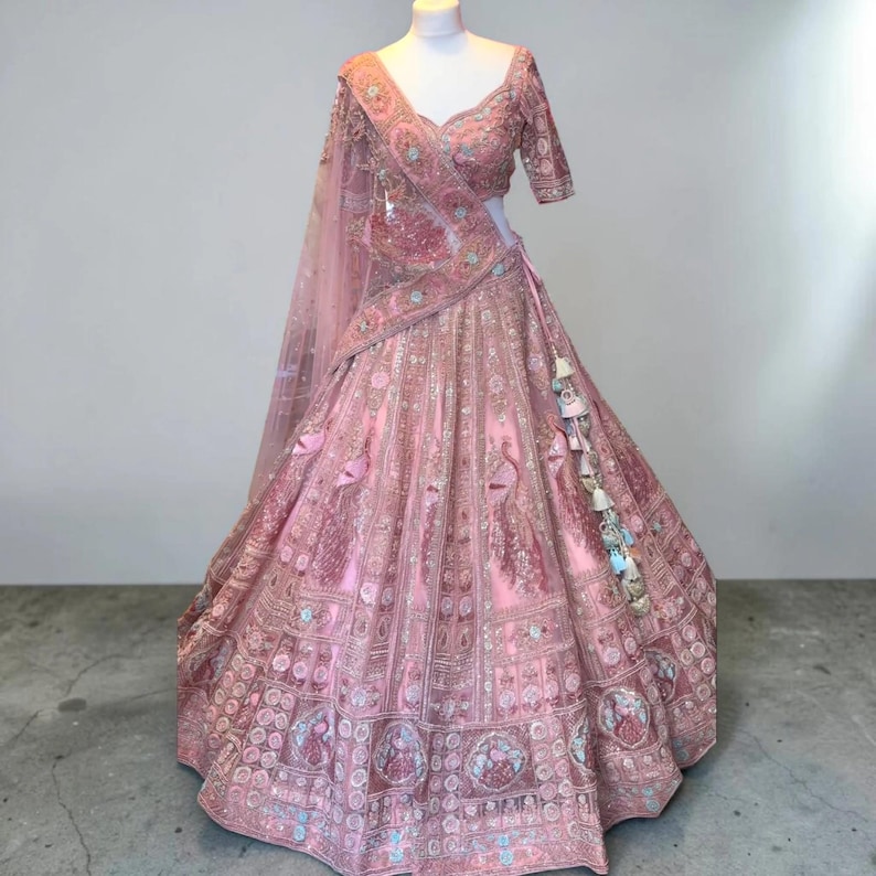 May include: A light pink lehenga choli set with detailed embroidery. The set comprises a short-sleeved top, a long skirt, and a sheer dupatta. The skirt and top are adorned with floral and peacock embroidery. Tassels are attached to the skirt.