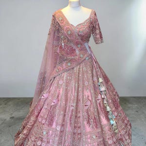 May include: A light pink lehenga choli, heavily embroidered with floral and peacock designs. The set includes a matching dupatta and decorative tassels. The outfit is displayed on a mannequin, showcasing the detailed craftsmanship.