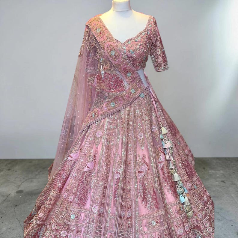 May include: A light pink lehenga choli, heavily embroidered with floral and peacock designs. The set includes a matching dupatta and decorative tassels. The outfit is displayed on a mannequin, showcasing the detailed craftsmanship.