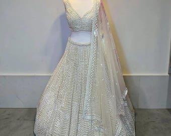May include: A white, intricately embroidered lehenga choli set on a mannequin. The set includes a sleeveless, V-neck top and a full, floor-length skirt. A sheer, embellished dupatta drapes over the mannequin. The fabric appears to have a subtle sheen.