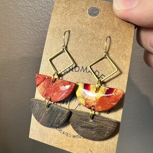 May include: A pair of handmade earrings with a geometric design. The earrings feature three tiers: a gold square, a red and orange semi-circle, and a brown wood semi-circle. The earrings are on a brown card with the text "Handmade" and "Created with Love".