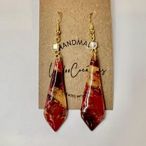 May include: A pair of red, gold and black teardrop-shaped earrings with gold hooks and clear crystal accents. The earrings are handmade and feature a marbled design.