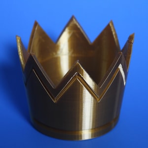 Peut inclure: Un pot de fleurs en plastique en forme de couronne de couleur dorée. Le pot a une finition lisse et brillante et est conçu pour contenir de petites plantes ou des succulentes.