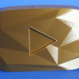 May include: A gold-coloured, geometric-patterned award plaque with a play button symbol. The plaque has a faceted surface and a raised border, set against a blue background.