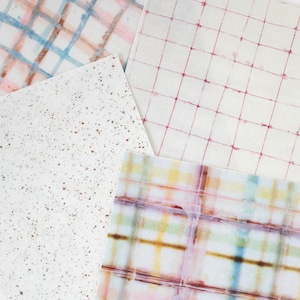 May include: An overhead shot of four pieces of paper with different patterns. One has a grid of red lines, another has a splatter design, and the other two have plaid patterns in various colors, including blue, pink, and yellow.