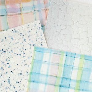 May include: A collection of watercolor paper sheets with various designs. One sheet features a grid pattern in pastel colors, another has a white cracked texture, and a third has blue paint splatters. The last sheet has a plaid pattern in blue, green, and pink.