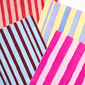 May include: An overhead shot displays four sheets of paper with colorful striped patterns. The sheets feature red, pink, blue, yellow, and pink stripes. The stripes are of varying widths and orientations, creating a vibrant and playful design.