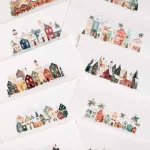 May include: A collection of watercolor greeting cards featuring various winter village scenes. The cards showcase colorful houses, shops, and festive decorations, with snow-covered rooftops and trees. The cards are arranged on a white surface.