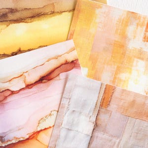 May include: An assortment of abstract art prints in warm, earthy tones. The prints feature watercolor-like textures with shades of brown, yellow, and pink. One print has a patchwork design, while others showcase fluid, layered patterns.