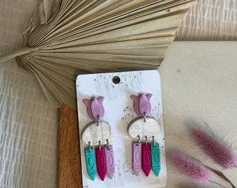 May include: A pair of dangle earrings with a pink flower charm, a white cloud charm, and three dangling charms in pink, teal, and red. The earrings are on a white card with a floral design.