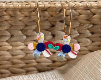 May include: A pair of gold hoop earrings with colorful flower-shaped charms. The charms are made of white clay with red, blue, yellow, green, and pink accents.