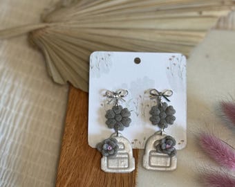 May include: A pair of silver backpack earrings with gray flower accents. The earrings are on a white card with a floral design.
